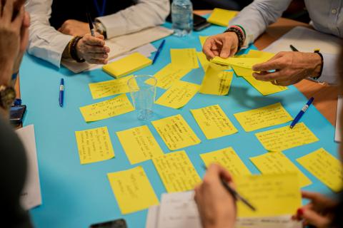 The image shows sticky notes on a table, surrounded by people's hands.